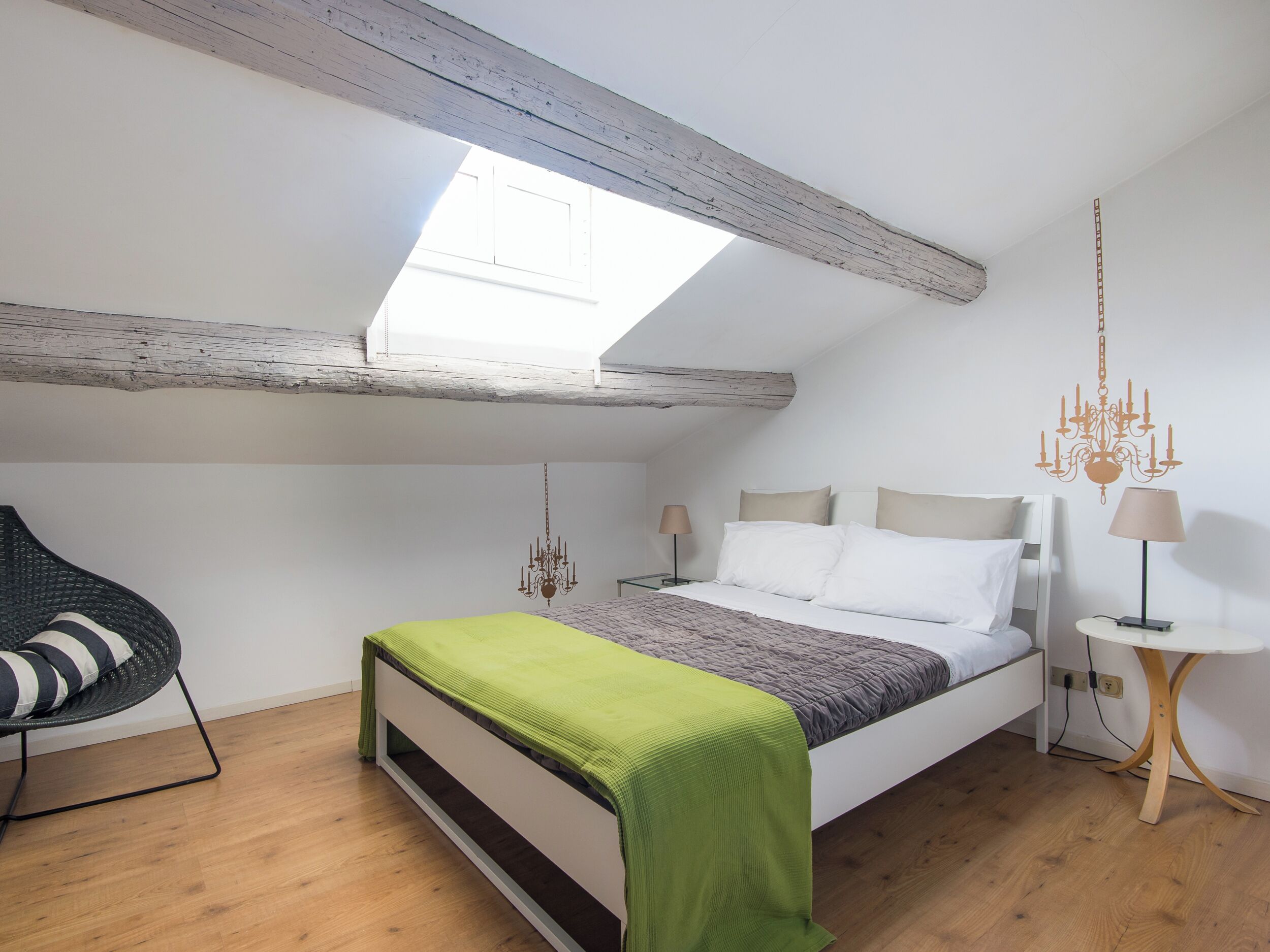 Ein Schlafzimmer mit schrägen Wänden, einem Bett mit grüner Decke, einer hängenden Chandelüster und einem kleinen runden Tisch mit Lampe.