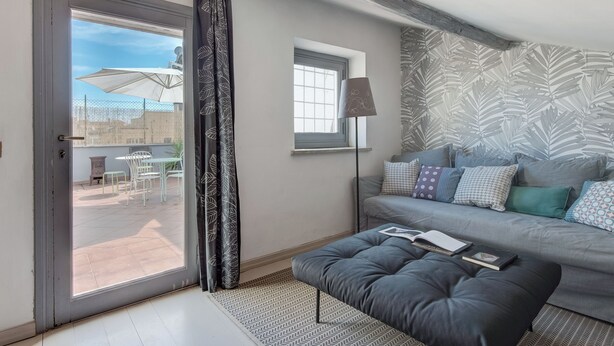 A modern living room with a grey sofa, patterned cushions, a black ottoman, and a floor lamp. A sliding glass door leads to an outdoor patio area with a table and chairs. - British Embassy Terrace Apartment (Rome)