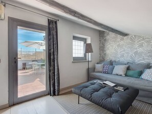 A modern living room with a grey sofa, patterned cushions, a black ottoman, and a floor lamp. A sliding glass door leads to an outdoor patio area with a table and chairs.