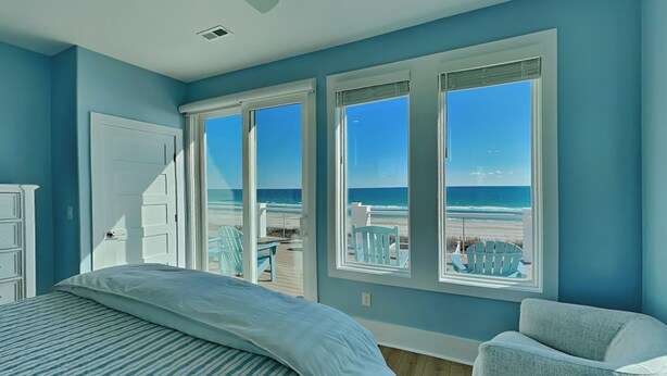 Un dormitorio con una cama grande, una silla y vistas a la playa a través de tres ventanas.