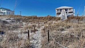 Una casa de playa con una escalera que lleva a una terraza, rodeada de arena y vegetación escasa.