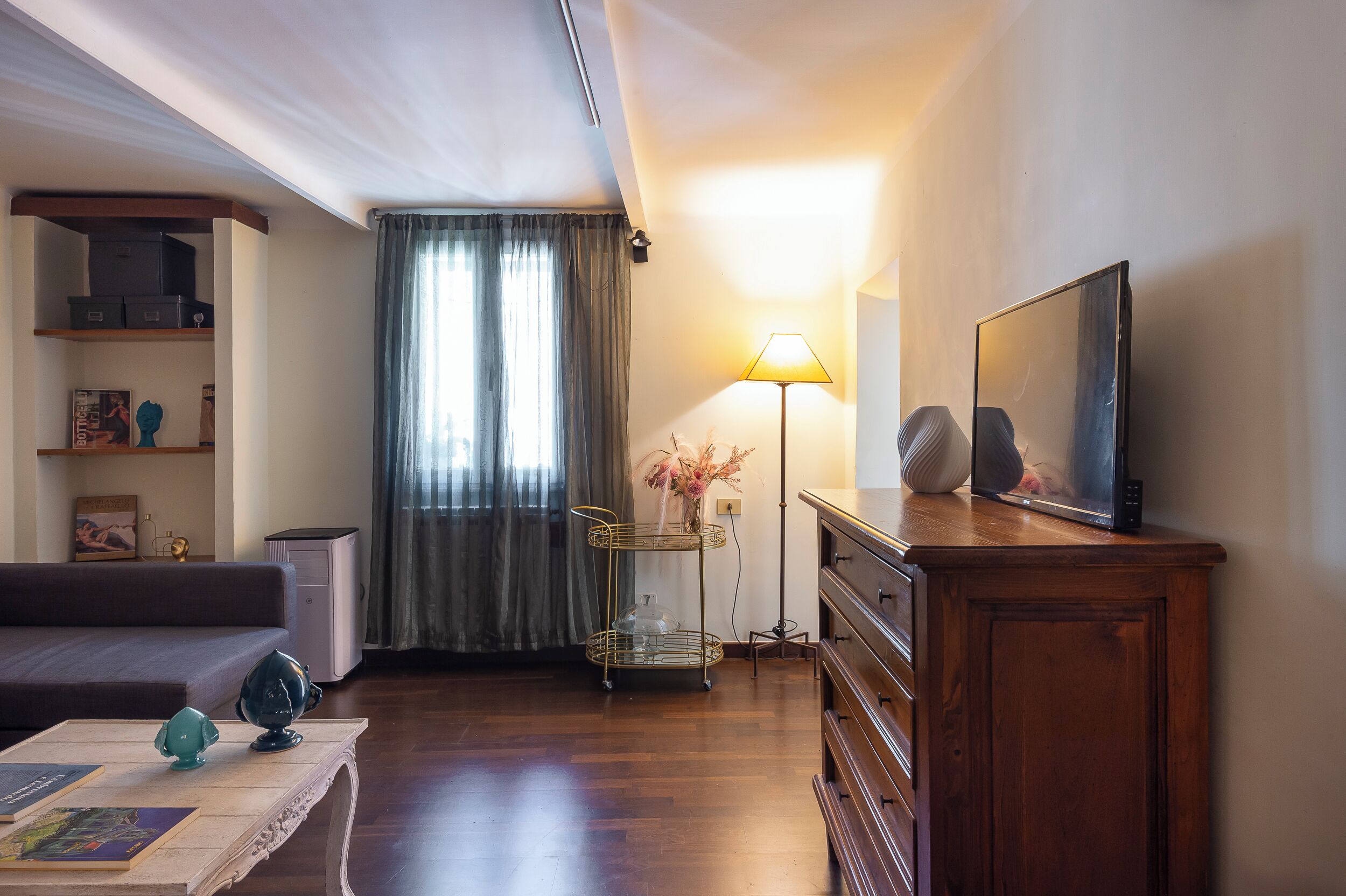 Ein Wohnzimmer mit einer Couch, einem Couchtisch mit Büchern, einem Fernseher auf einer Holzkommode, einer Lampe und einem Fenster mit Vorhängen.