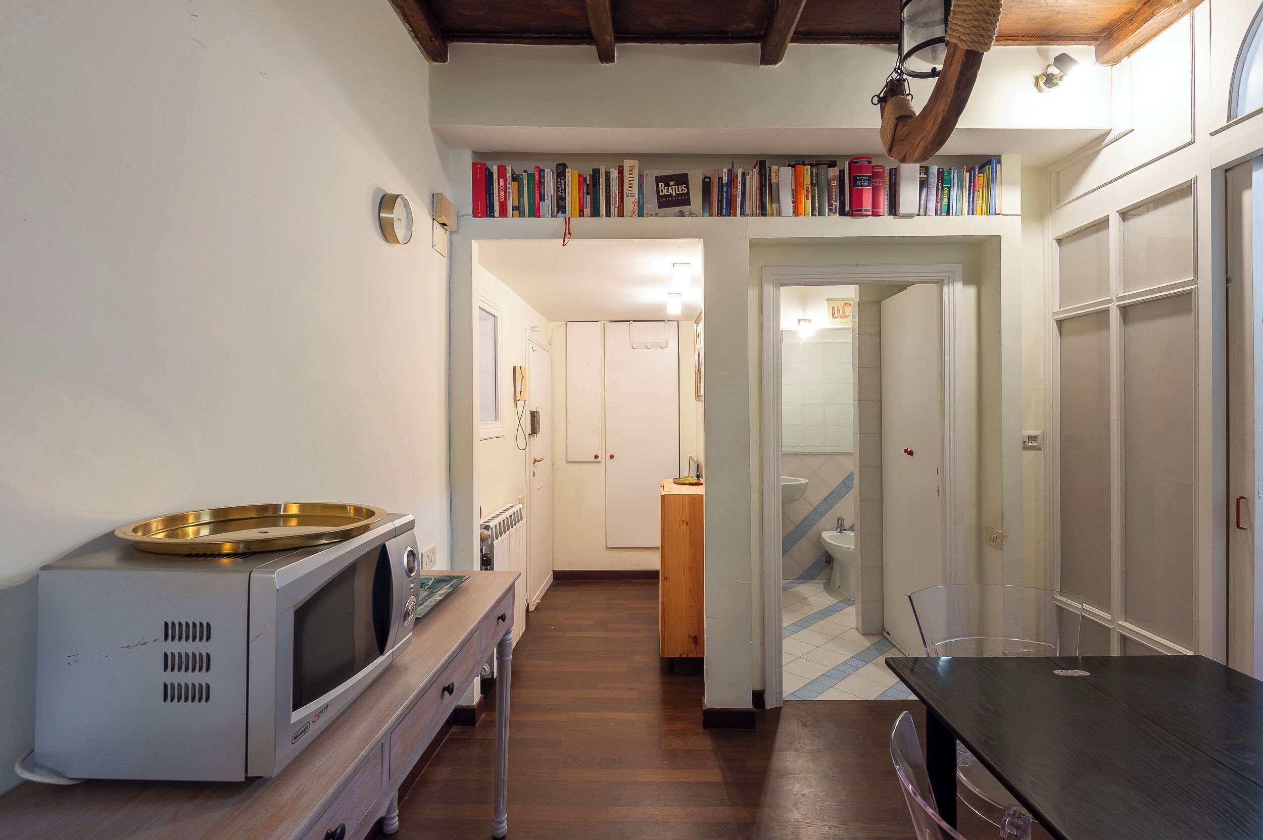 A compact kitchen with a microwave, a wooden table, and a bookshelf above the counter.