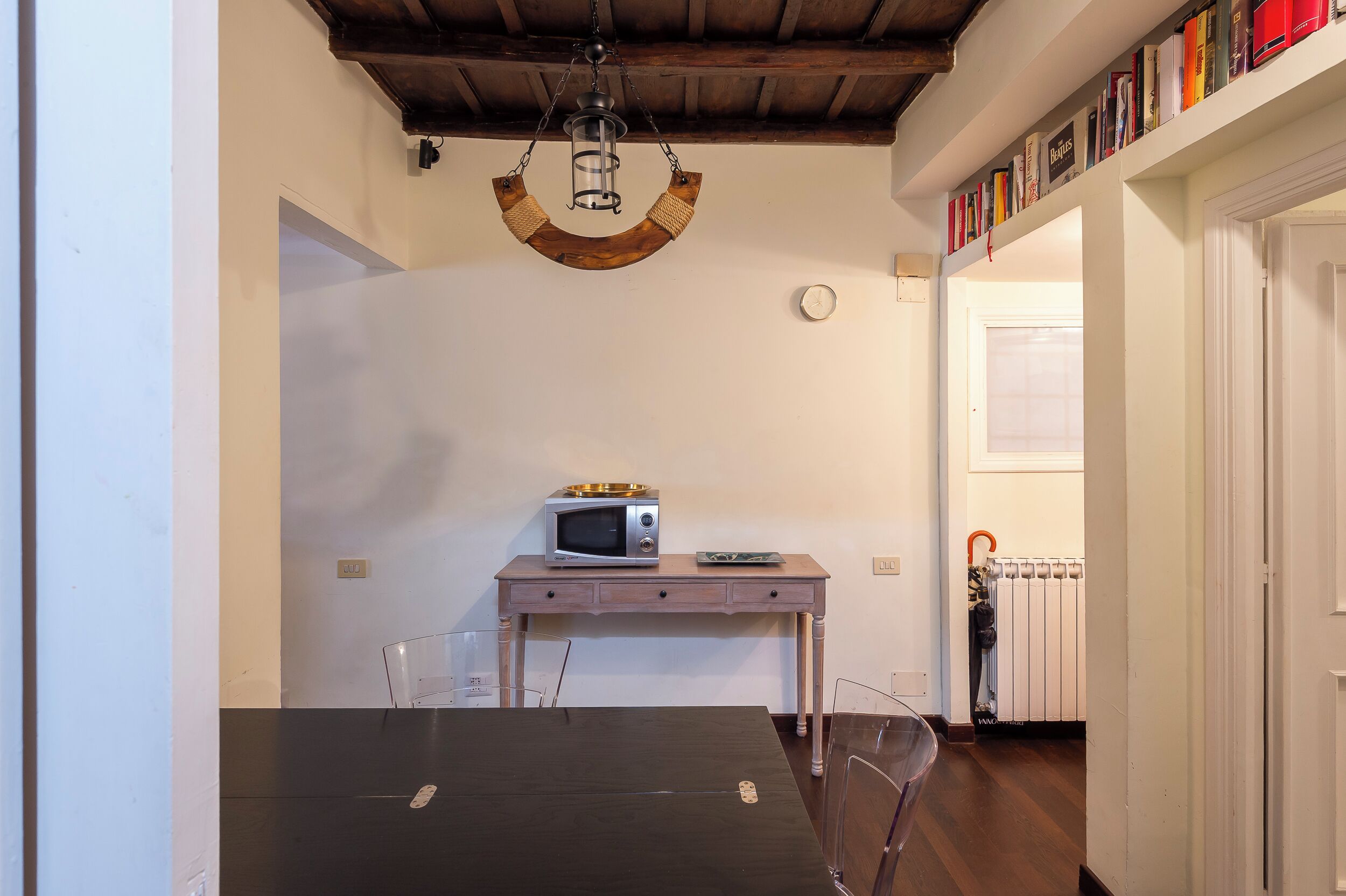 A dining area with a wooden table, chairs, a microwave, and a small wooden desk with a lamp.