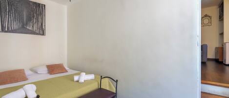 Une chambre d’hôtel avec deux lits, l’un recouvert d’une couvre-lit verte et l’autre d’une couvre-lit marron. Un tableau est accroché au mur au-dessus des lits, et un escalier mène à une autre partie de l’hôtel.