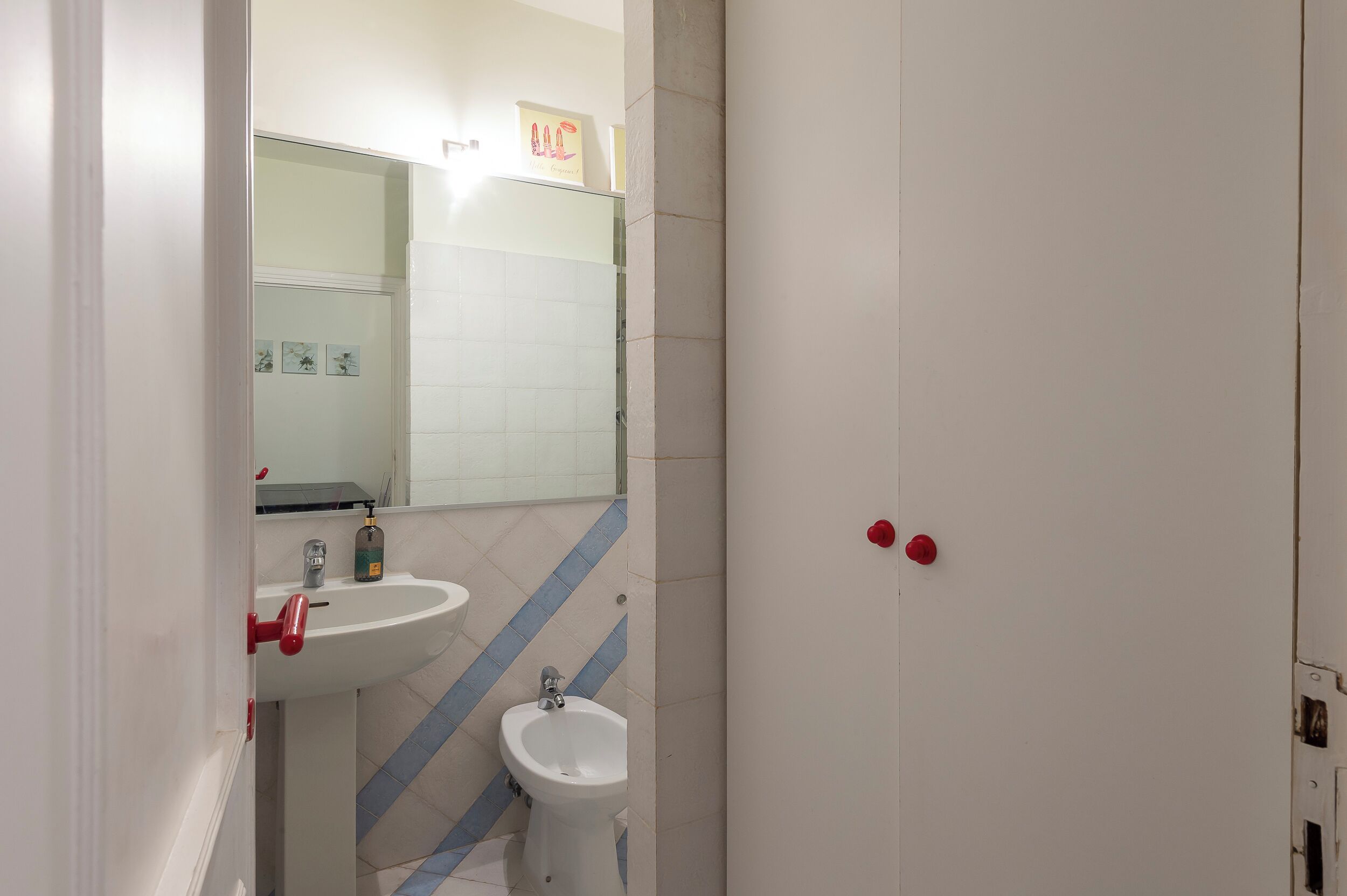 A bathroom with a white sink, a mirror, and a bidet. There are two red handles on the wall.