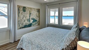 Ett sovrum med en stor säng, en takfläkt och utsikt över stranden genom fönstren.