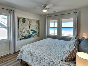 A bedroom with a large bed, a ceiling fan, and a beach view through the windows. - Walking on Sunshine by Topsail Realty (Holly Ridge)