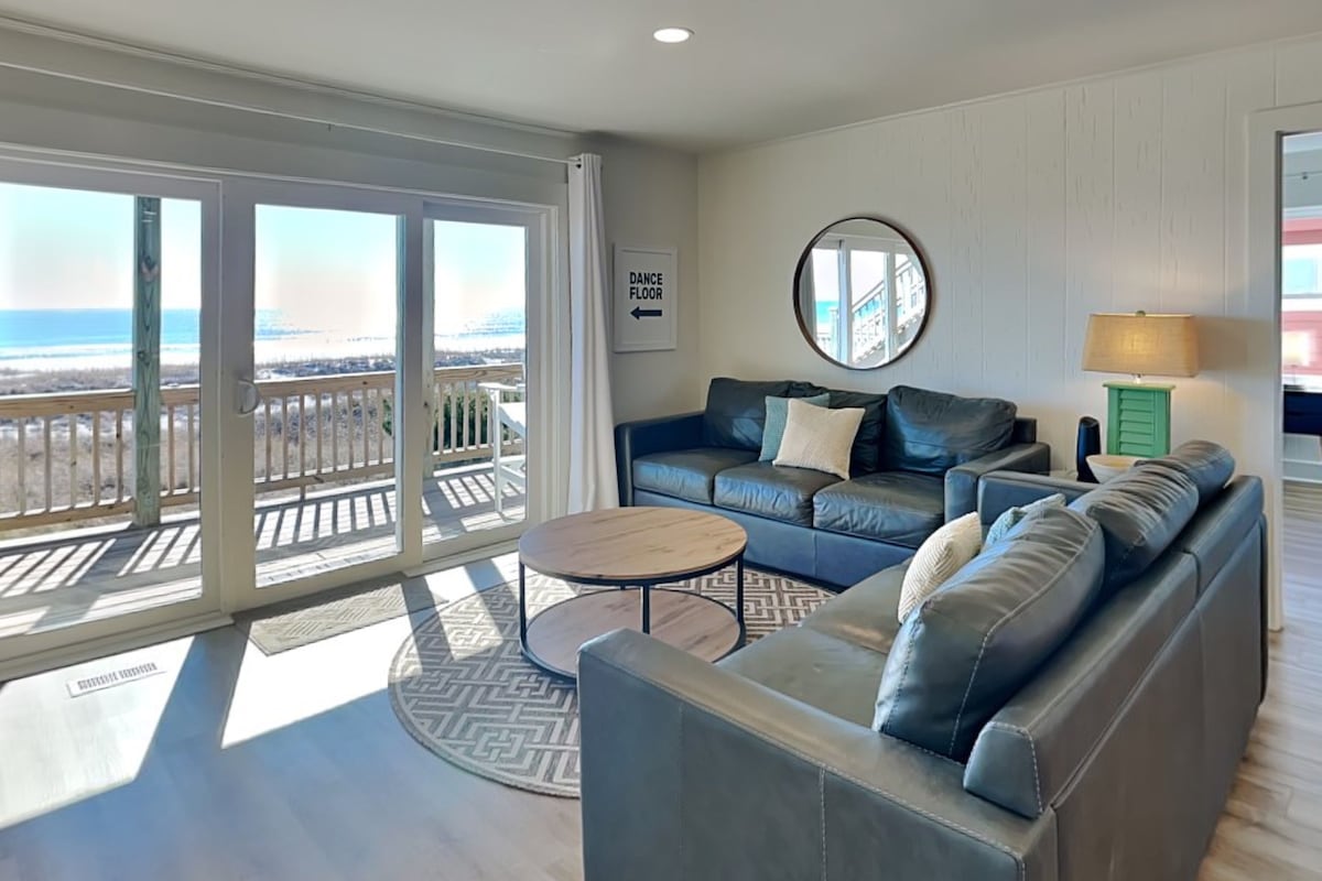 A spacious living room with a large sliding glass door leading to a balcony with a view of the beach.