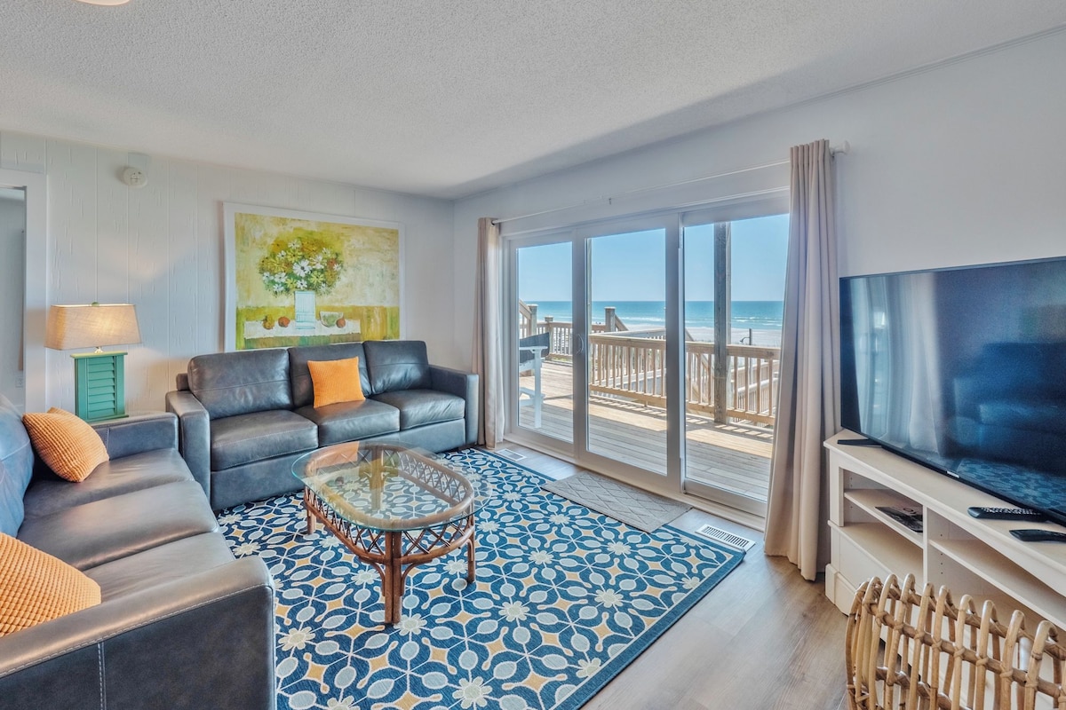 A living room with a sofa, coffee table, and TV, overlooking a beach.