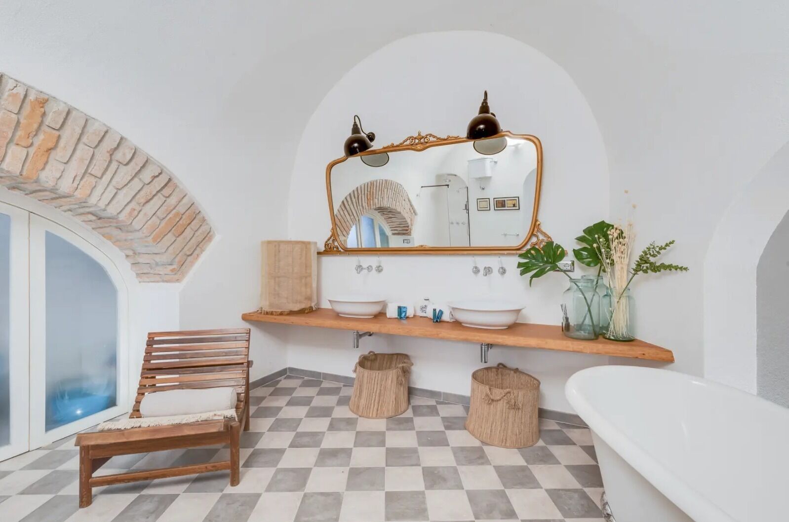 A bathroom with a checkered floor, wooden bench, two sinks, a large mirror, and a bathtub.