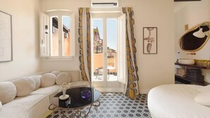 Ett hotellrum med en böjd soffa, ett runt bord, en säng, ett stort fönster med gardiner och utsikt över byggnader utanför.