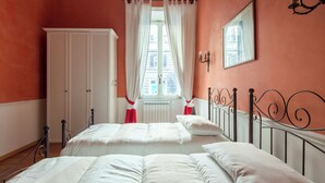 Une chambre à coucher avec un lit, des rideaux blancs, un radiateur, une armoire et une fenêtre donnant sur des immeubles.