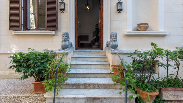 L’entrée d’un bâtiment est agrémentée d’un escalier en pierre menant à une porte, de part et d’autre de laquelle se trouvent deux statues et des plantes en pot.