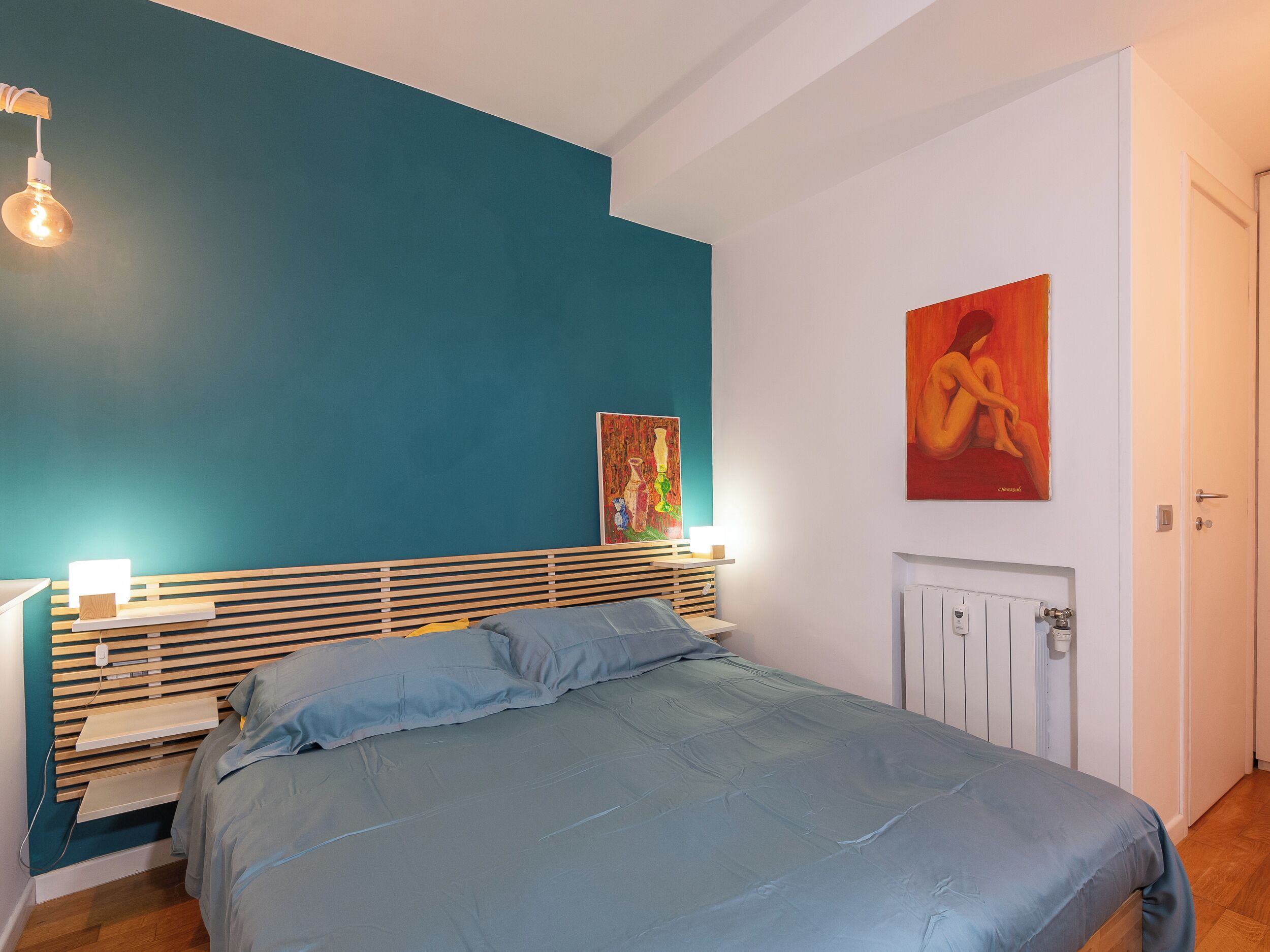 Quarto com parede de destaque azul-turquesa, cama com roupa de cama azul, cabeceira de madeira e duas luminárias de cabeceira.