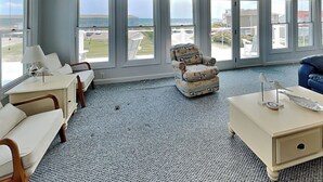 Um quarto espaçoso com janelas amplas, uma poltrona branca, uma mesinha de centro com abajur e um sofá azul.
