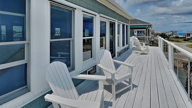 Um deck de madeira com cadeiras Adirondack brancas e grandes janelas.