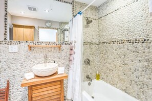 Un baño con pared de piedra, tocador de madera, lavabo redondo, bañera y cortina de ducha.