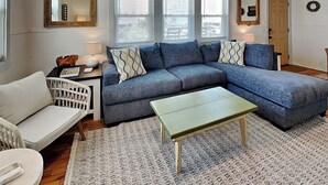 A living room with a sectional sofa, a small table, and a white armchair. There are three windows with blinds, a door, and a wall-mounted mirror. - Mountains to Sea by Topsail Realty (Holly Ridge)