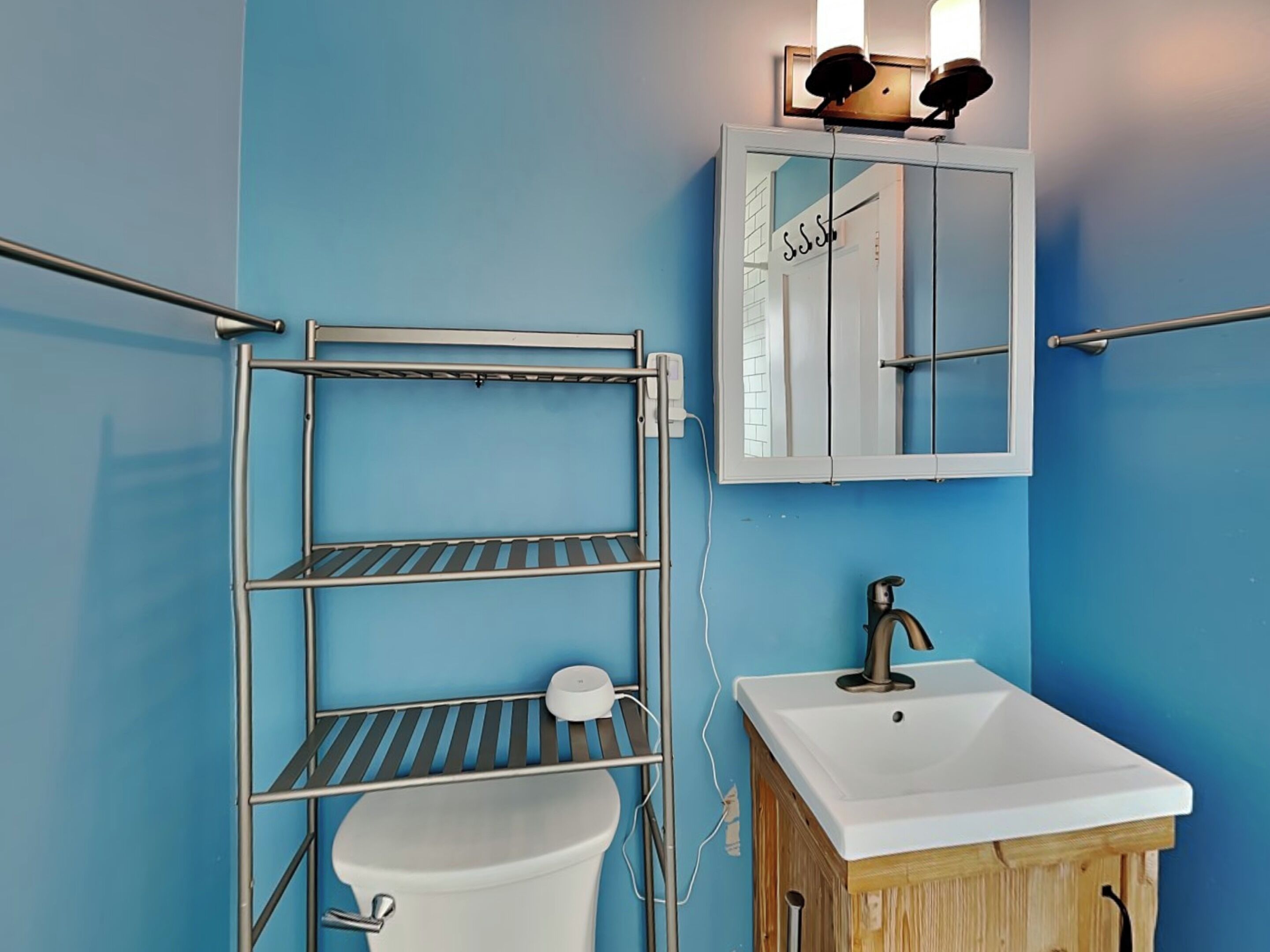 A bathroom with a blue wall, a white sink, a mirror, and a metal shelf unit.