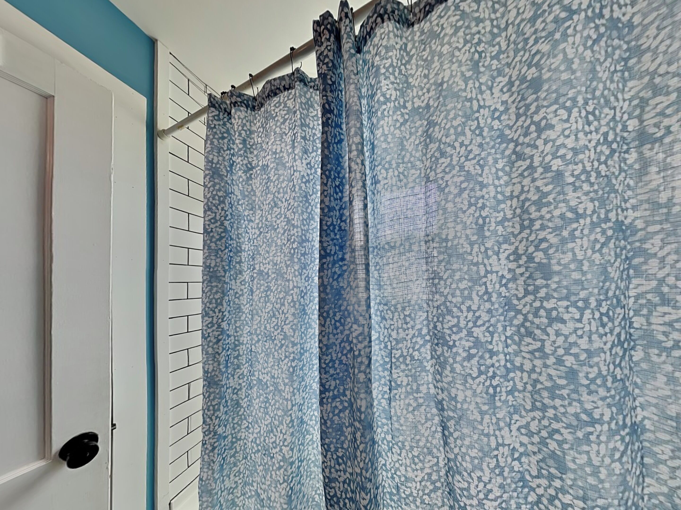 A bathroom with a white door, a white tiled wall, and a shower curtain with a blue and white leaf pattern.