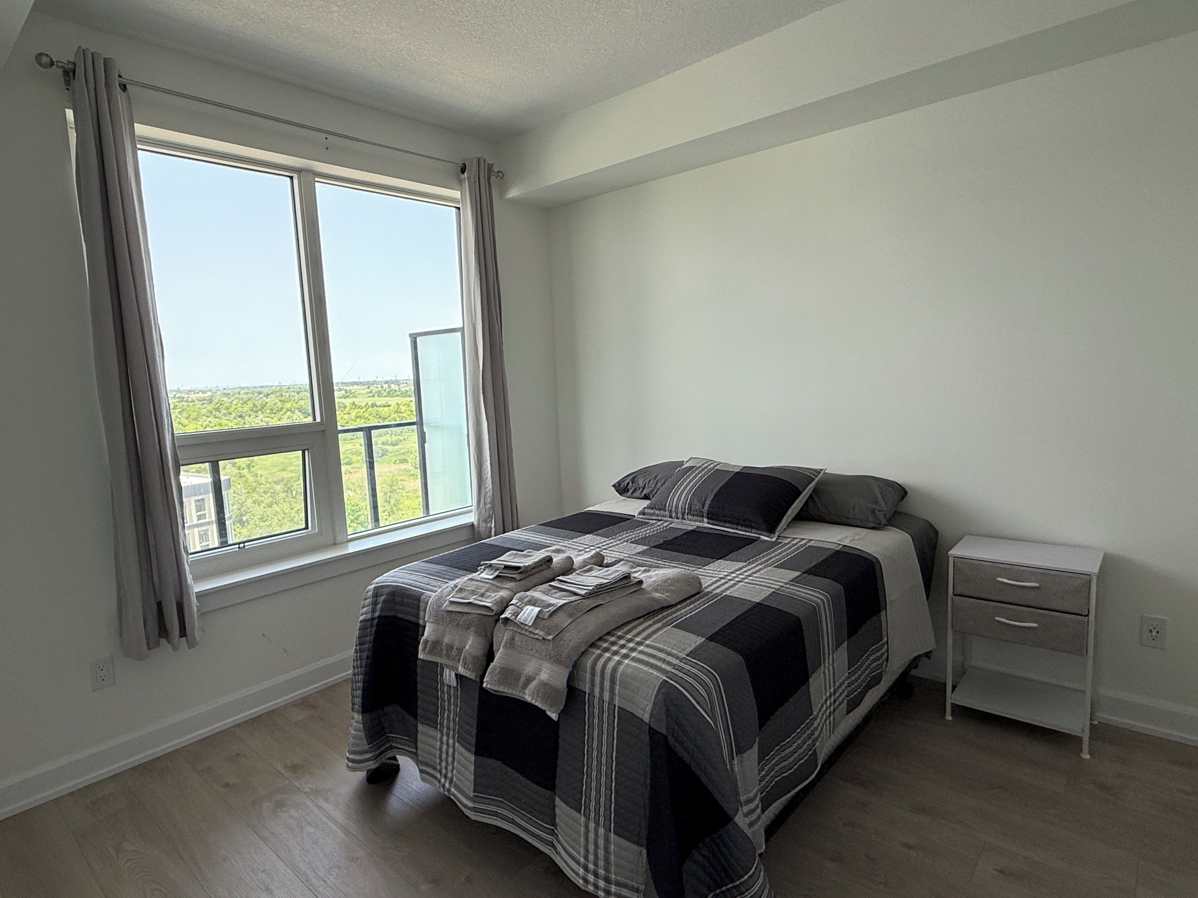 Een slaapkamer met een groot raam, een bed met een geruit sprei, een nachtkastje en een bedtafel.