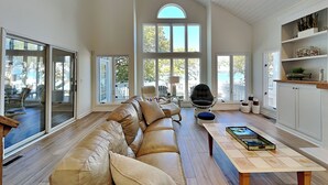 A spacious living room with a large leather sofa, a wooden coffee table, and a view of the outdoors through large windows. - The Pack House by Topsail Realty (Holly Ridge)