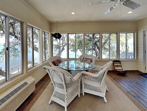 A sunlit room with a round glass table surrounded by white wicker chairs, large windows, and a ceiling fan. - The Pack House by Topsail Realty (Holly Ridge)