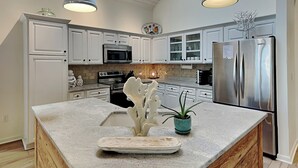 A kitchen with white cabinets, a stainless steel refrigerator, a microwave, and a wooden island with a decorative plant. - The Pack House by Topsail Realty (Holly Ridge)