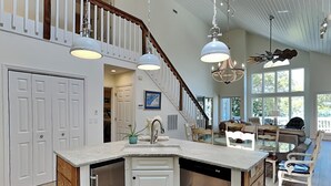 A spacious kitchen with a central island, white cabinetry, and a dishwasher. A staircase leads to a loft area with a dining space. - The Pack House by Topsail Realty (Holly Ridge)