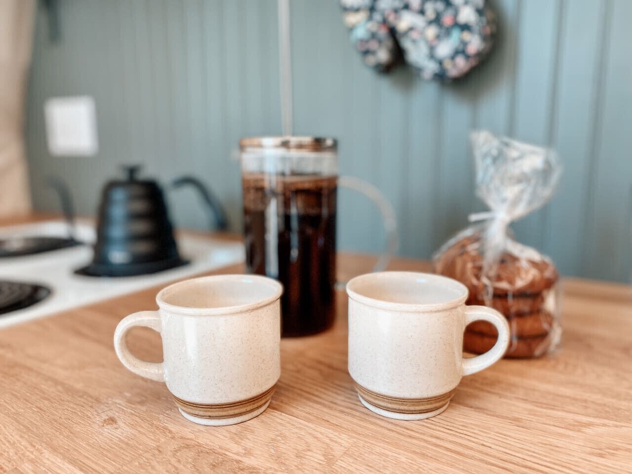 Deux tasses en céramique blanche sont posées sur une table en bois, avec une machine à café et un bocal en verre en arrière-plan.