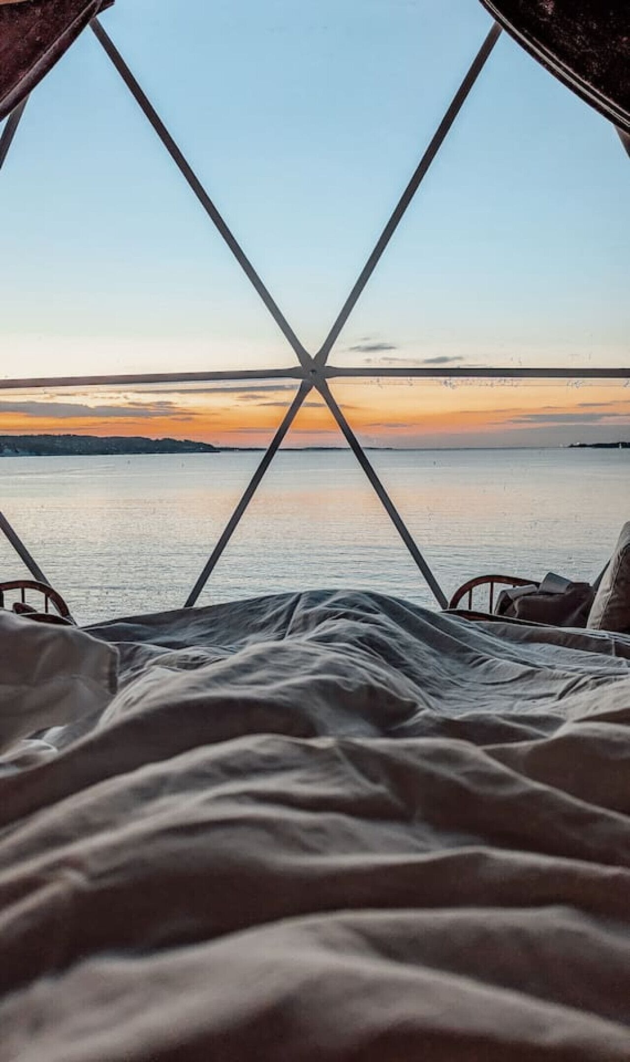 Un lit donnant sur un coucher de soleil sur une étendue d’eau, le tout encadré par une structure de fenêtre aux lignes géométriques.