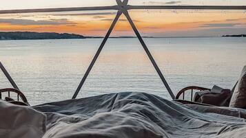 Un lit donnant sur un coucher de soleil sur une étendue d’eau, le tout encadré par une structure de fenêtre aux lignes géométriques.