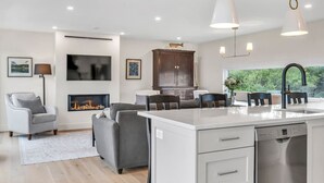 Een moderne keuken met een centraal eiland, een eethoek met stoelen, een open haard en uitzicht op groen.