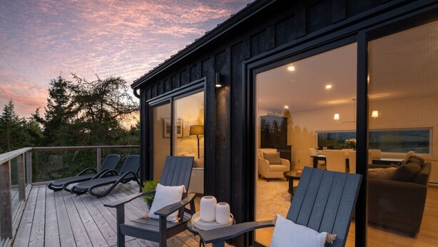 Een modern terras buiten met lounge stoelen en een tafel, met uitzicht op een goed verlichte binnenruimte met een bank en een televisie.