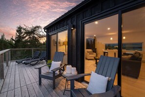 Une terrasse extérieure moderne, dotée de chaises longues et d’une table, qui offre une vue sur un espace intérieur bien éclairé, comprenant un canapé et un téléviseur.
