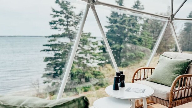 A cozy outdoor seating area with wicker furniture, a small round table, and binoculars, overlooking a forested landscape and a body of water. - Luxury Coastal Dome - Private Hot Tub, Oceanfront Deck, Firepit & Parking (LaHave)