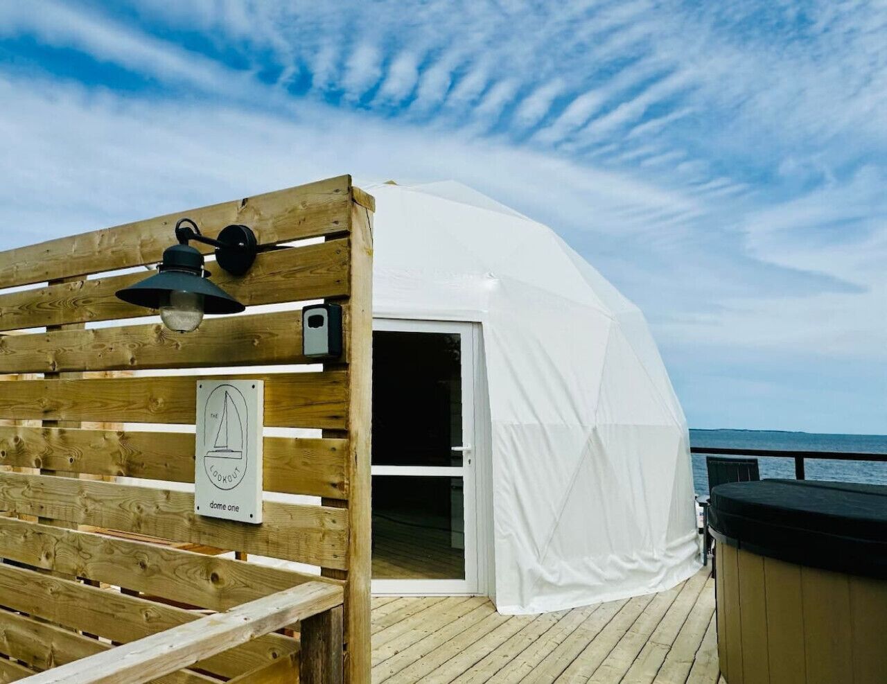 Eine Holzterrasse mit einer weißen Kuppelstruktur, einem Schild mit der Aufschrift 'Kuppel eins' und Blick aufs Meer.