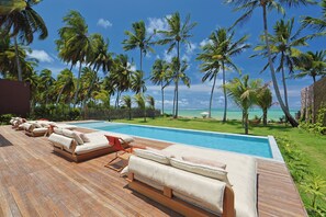 A wooden deck with lounge chairs and a pool, surrounded by palm trees and a view of the ocean. - Origem Patacho Design Hotel (Porto de Pedras)