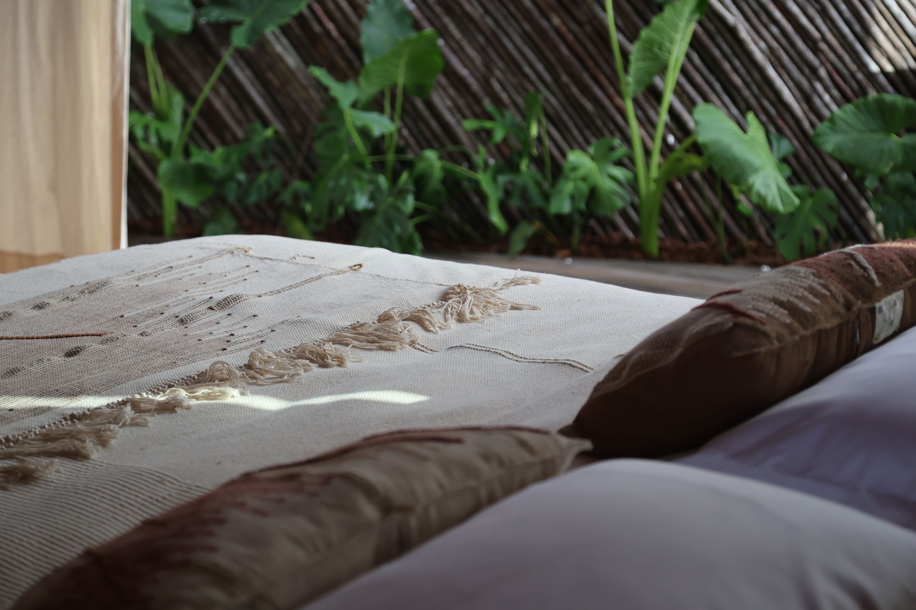 Un lit recouvert d’un drap blanc texturé et agrémenté d’un oreiller marron, le tout mis en valeur par un décor de plantes vertes et une clôture en bambou.