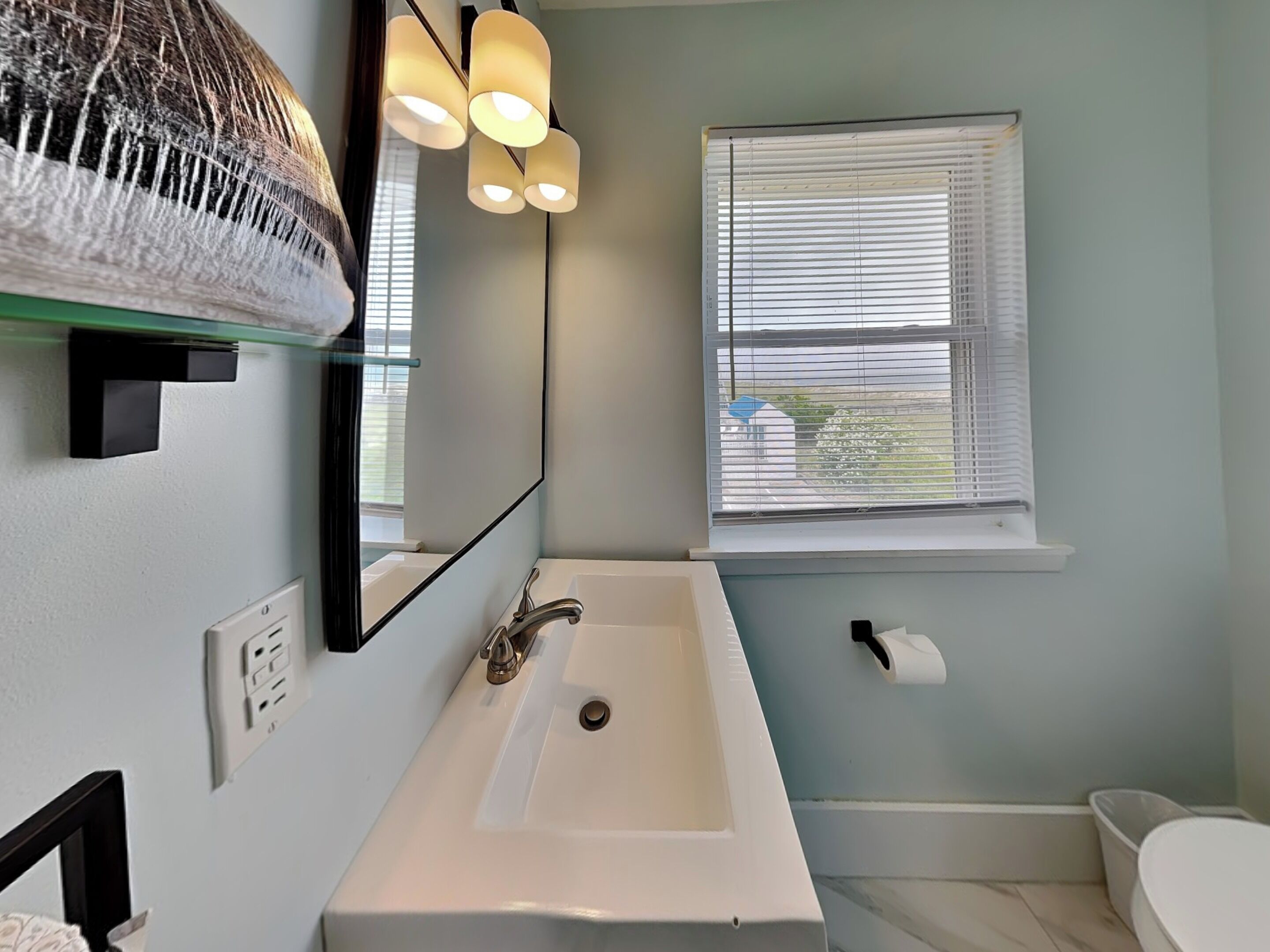 Ein Badezimmer mit einem weißen Waschbecken, einem Spiegel, einem Handtuchhalter, einem Fenster mit Jalousien und einer Toilette.