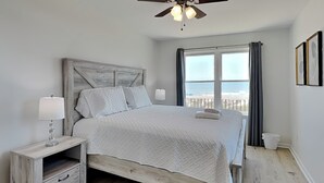 A bedroom with a bed, a nightstand, a ceiling fan, a window with curtains, and a view of the beach. - Seaside Serenity by Carolina Retreats (Carolina Beach)