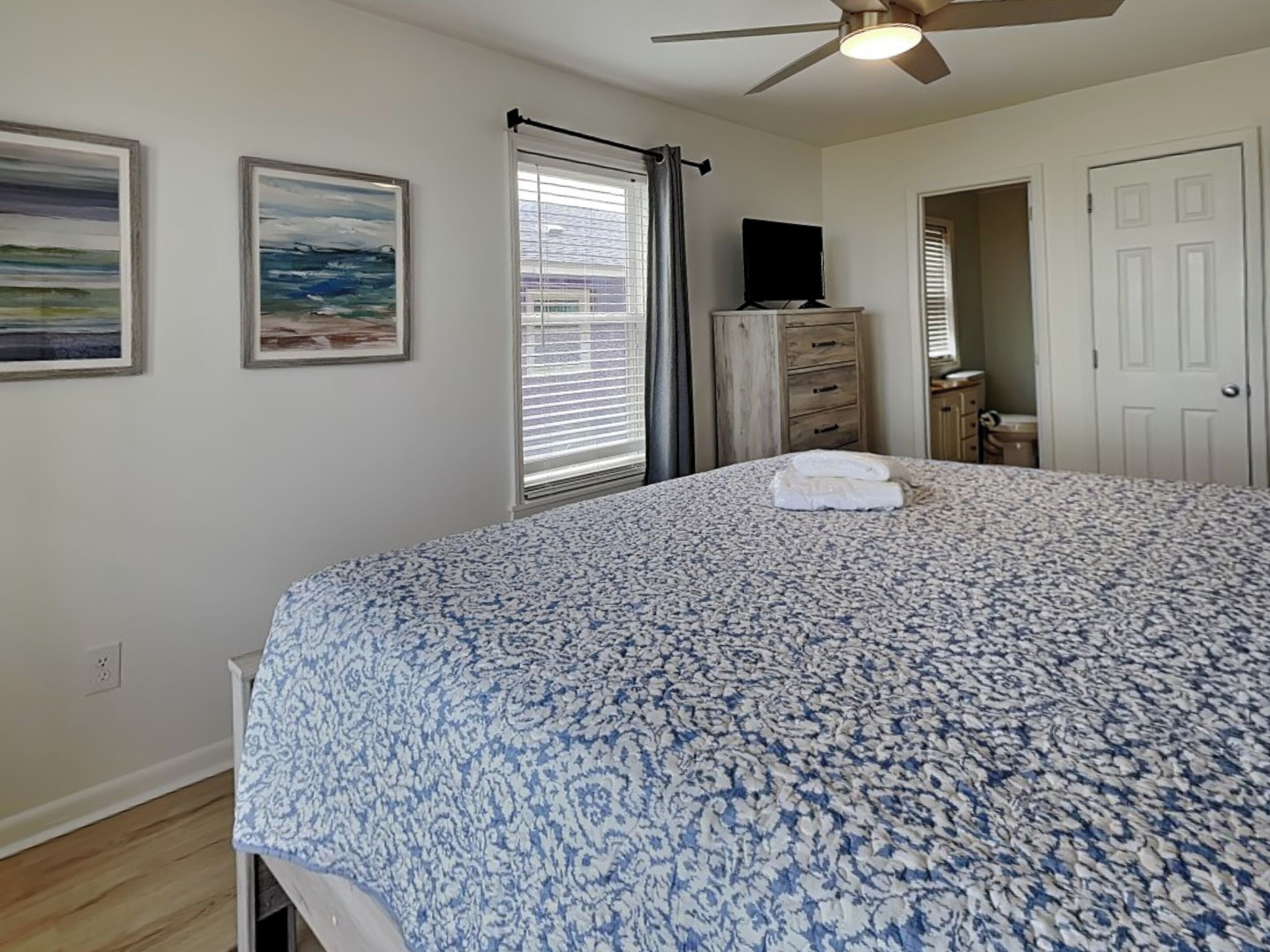 A bedroom with a large bed, a ceiling fan, a dresser, and two framed paintings on the wall.