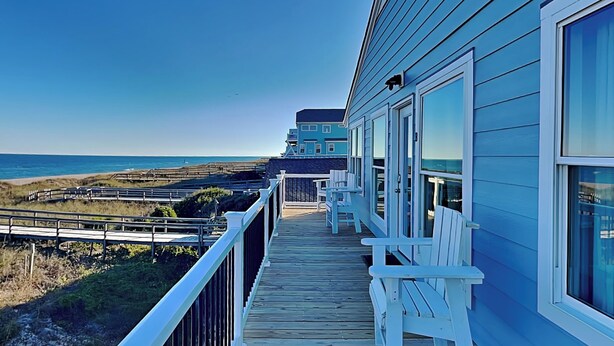 A wooden deck with white chairs, overlooking a beach and a building with large windows. - Seaside Serenity by Carolina Retreats (Carolina Beach)
