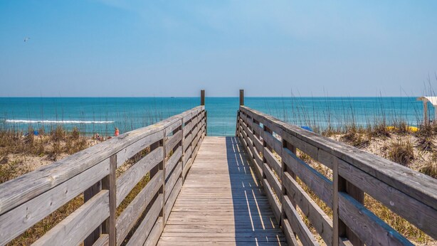 A wooden boardwalk leading to a beach with clear blue water and a clear sky. - The Sandcastle @ Tiki by Carolina Retreats (Carolina Beach)