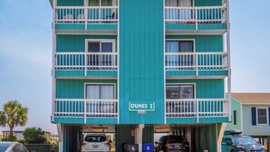 A multi-story building with a central vertical strip, balconies on each floor, and a street sign reading "DUNES I 1225". - The Sandcastle @ Tiki by Carolina Retreats (Carolina Beach)