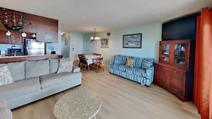 A living room with a sofa, a dining table, a cabinet, and a small round table. - The Sandcastle @ Tiki by Carolina Retreats (Carolina Beach)