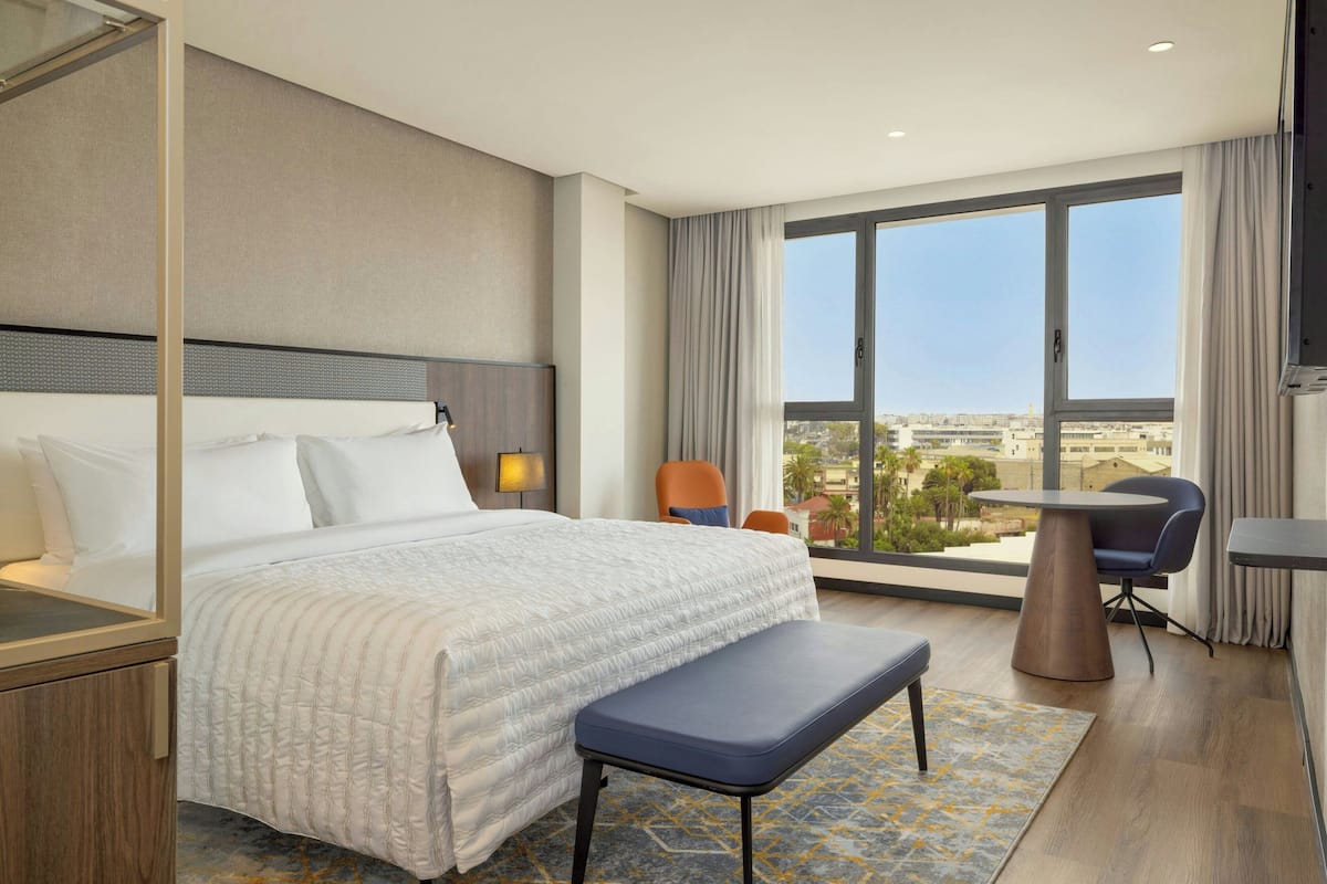 Habitación de hotel con una cama grande, un banco, una mesita y vistas a la ciudad a través de amplios ventanales.
