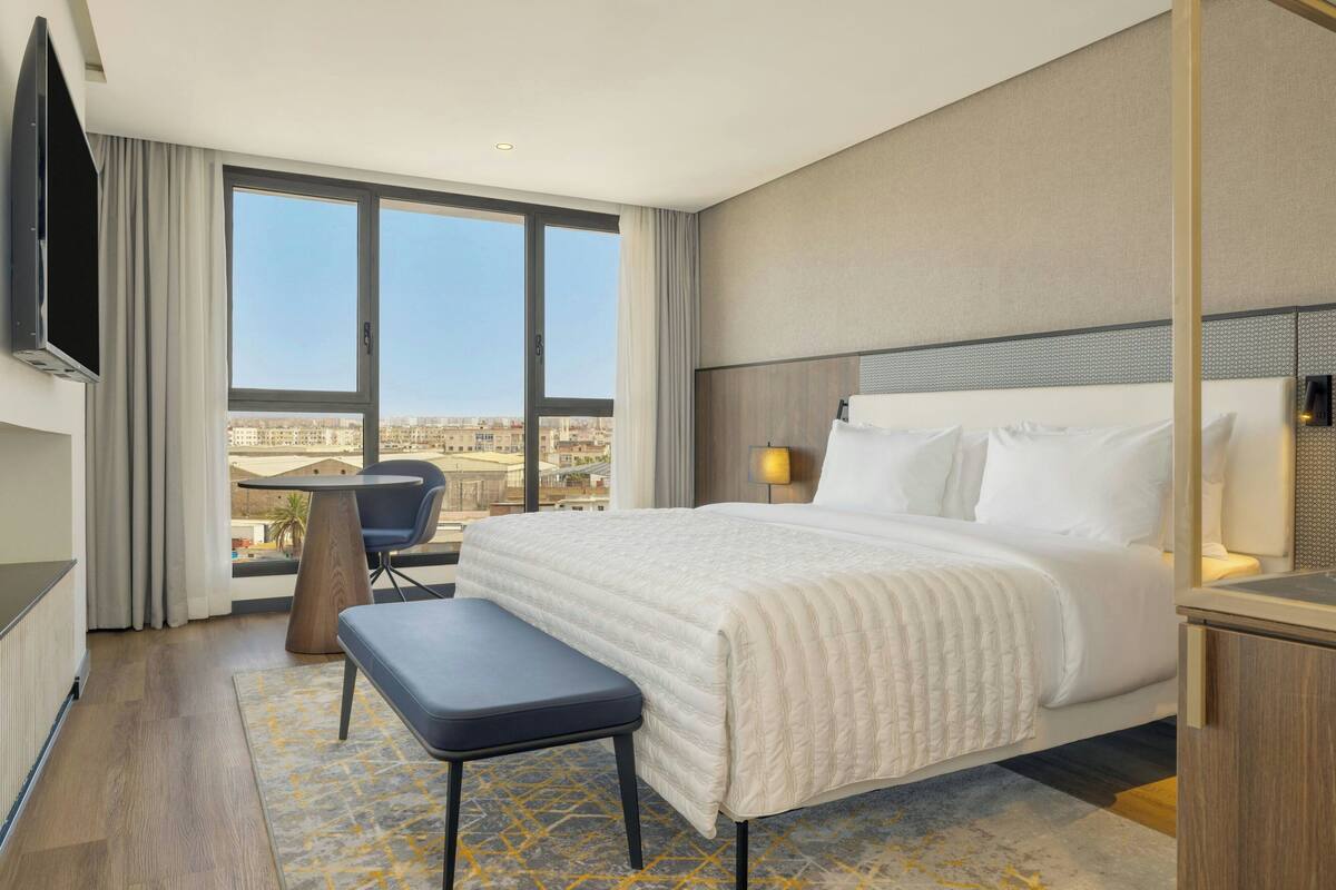 Habitación de hotel con una cama grande, un escritorio, una silla, un televisor y vistas a la ciudad.