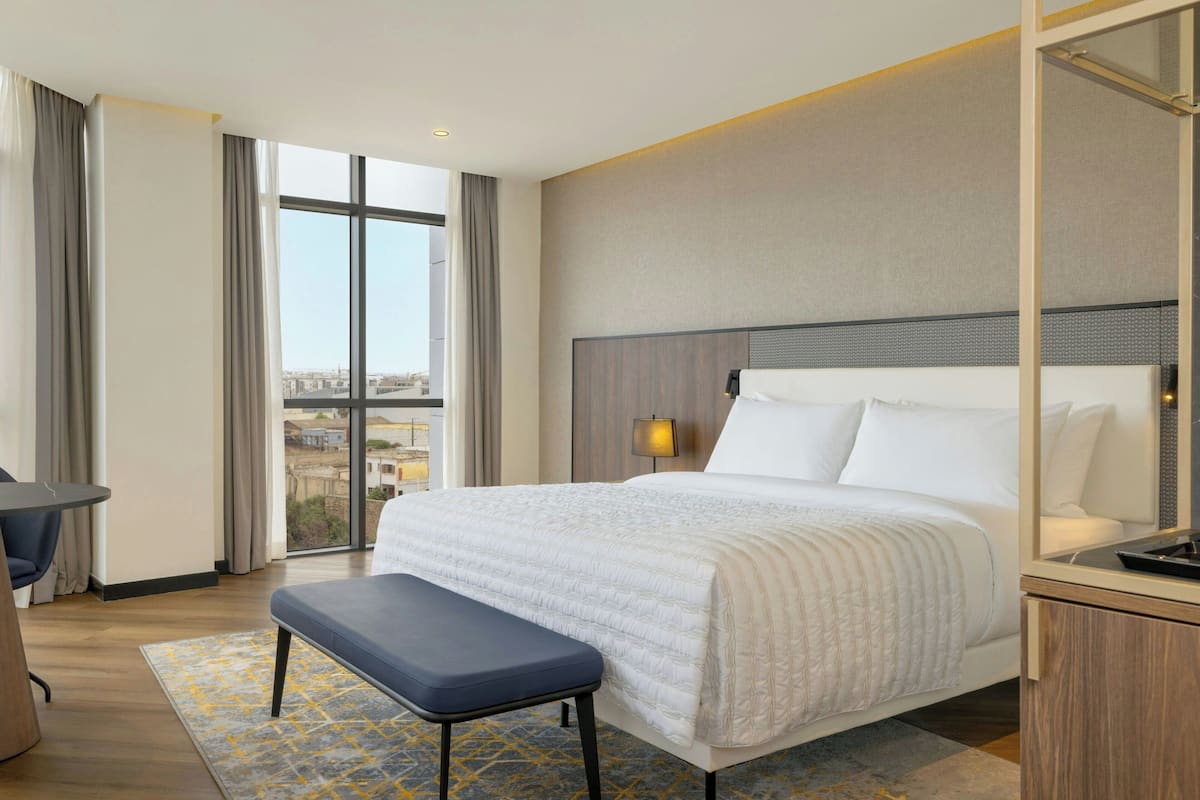 Habitación de hotel con una cama grande, una banca, un escritorio y vista a la ciudad a través de amplias ventanas.